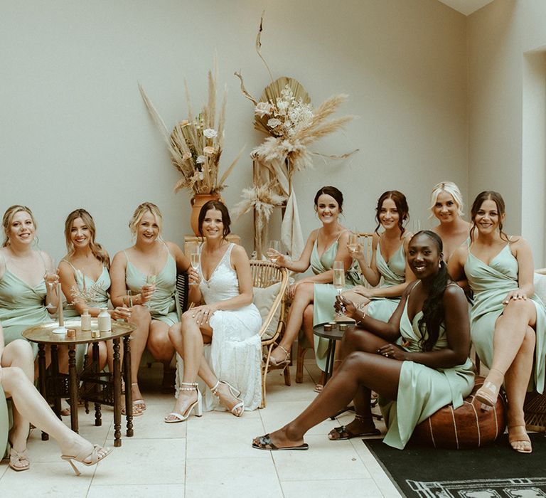 Bride in lace wedding dress sits with her ten bridesmaids in sage green satin wrap dresses before the wedding with dried flower decor