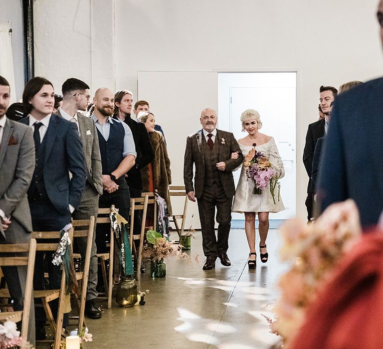 Father of the bride in checkered three piece suit walks the bride in a short wedding dress down the aisle