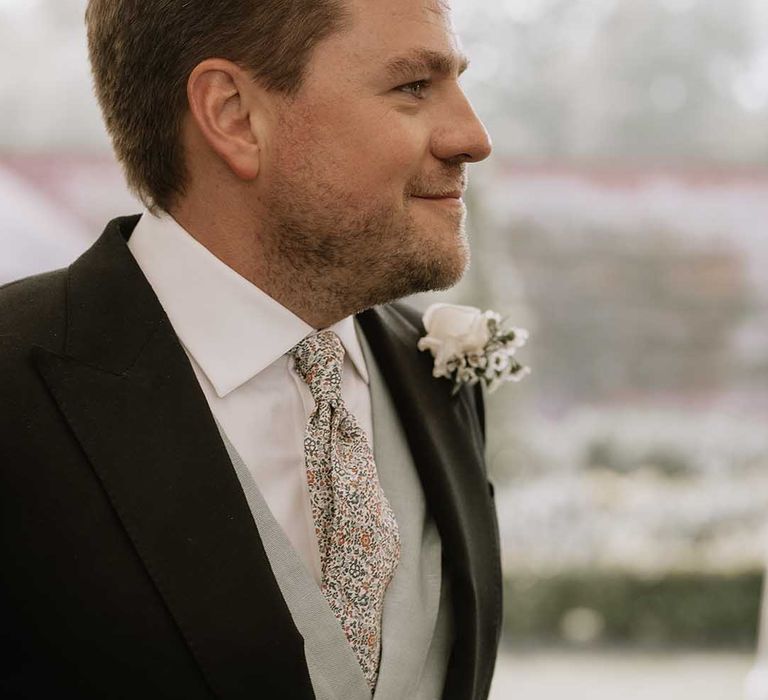 Groom in a green waistcoat and floral patterned tie and white flower buttonhole smiles as he sees the bride for the first time 