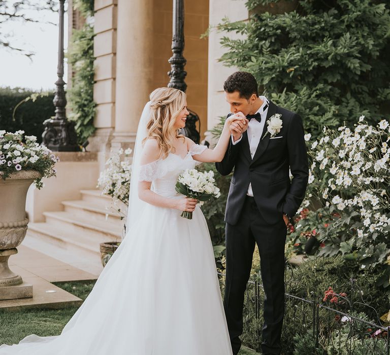 Groom in black tie kisses the bride's hands as she wear an off the shoulder wedding dress and white flower bouquet 