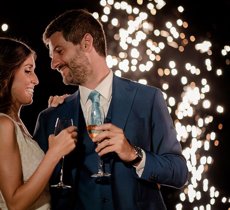 Bride & groom look lovingly at one another outdoors on their wedding day as sparklers light behind them 