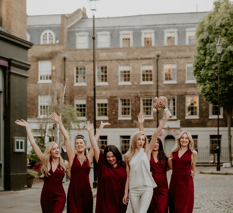 Bride in second wedding outfit with top and trousers walking the streets of London with her bridal party in dark red bridesmaid dresses 