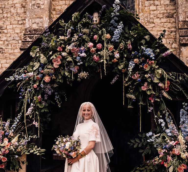 Bride in lace top Jesus Piero wedding dress standing in front of the church decorated with pink and blue wedding flowers 