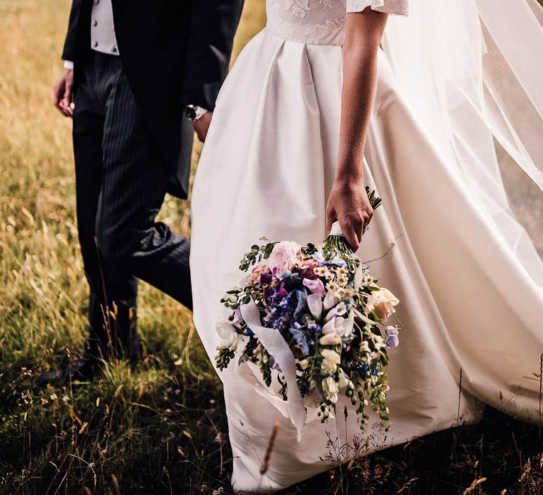 Smiling bride and groom walk through a field together as the bride carries her colourful bridal bouquet 