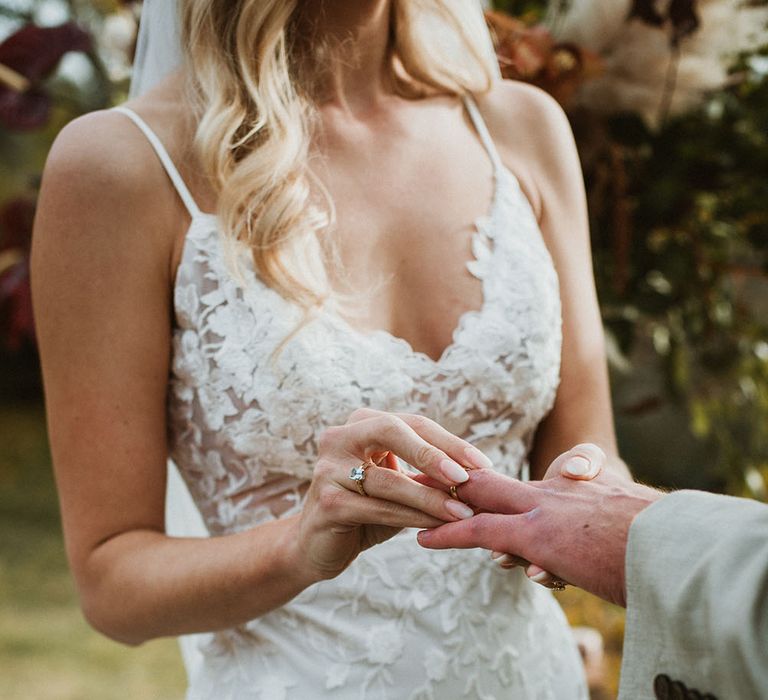 Bride places wedding ring on her groom during wedding ceremony and wears Emmy Mae Bridal lace wedding dress with thin straps and low neck