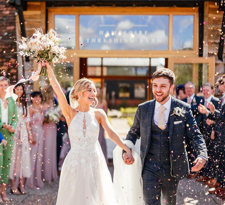 Bride in halter neck wedding dress lifts her pink and white bouquet into the air walking hand in hand with the groom 