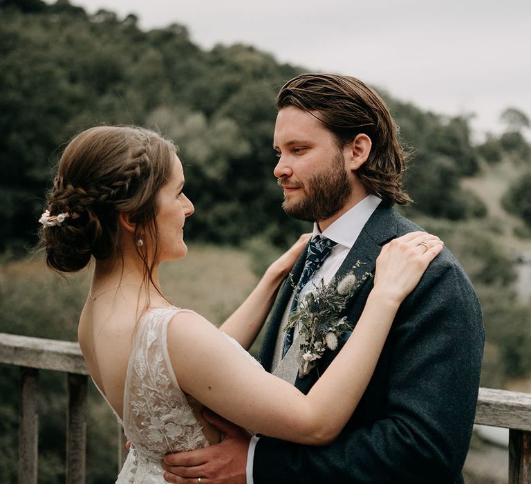 Bride wears her brown hair in low bun with plaits either side whilst she looks lovingly at her groom 