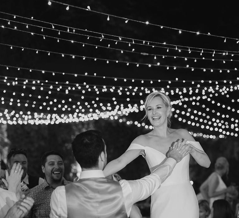 Bride dances on chair during outdoor wedding with her groom as fairy light canopy hangs above them