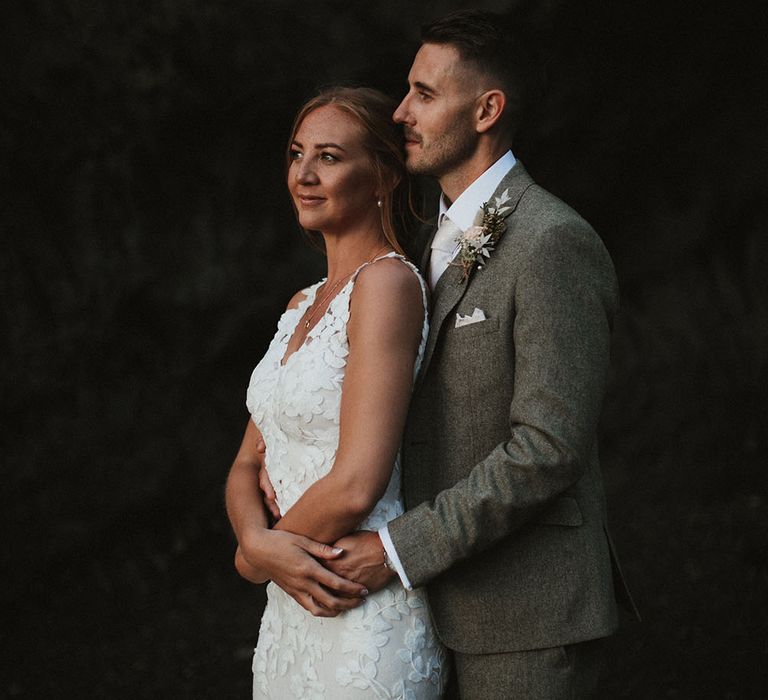 Bride wears fitted floral embellished Enzoani wedding dress as her groom stands behind her wearing green suit with white pocket square and floral buttonhole