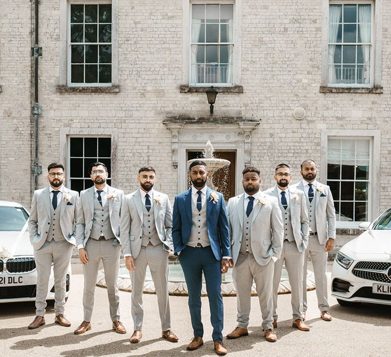 Groom in three piece blue suit with grey waistcoat stands with his groomsmen in grey three piece suits 