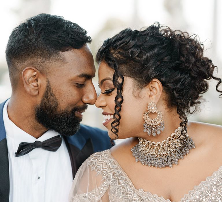 Bride wears her black hair in up-do with tight curls to the front of her face and statement jewellery 