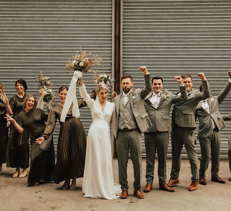 Bridesmaids in green bridesmaid dresses and groomsmen in grey suits stand with bride in Reformation wedding dress and groom in three piece suit 