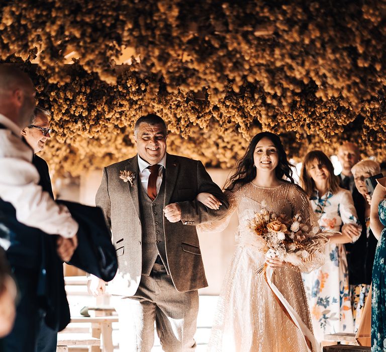 Bride in long sleeve gold wedding dress walks with the aisle with her father as dried foliage hangs above them