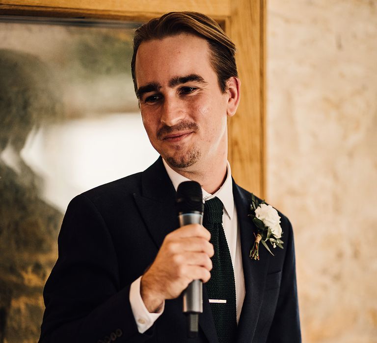 Groom stands with a microphone to deliver a speech in a navy blue suit and white rose buttonhole 