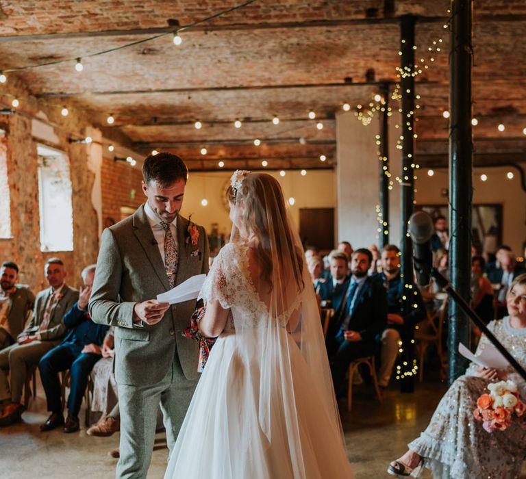 Groom in a grey tweed suit with flower tie standing at the altar with the bride in a tulle and lace wedding dress for their hand fasting ceremony 