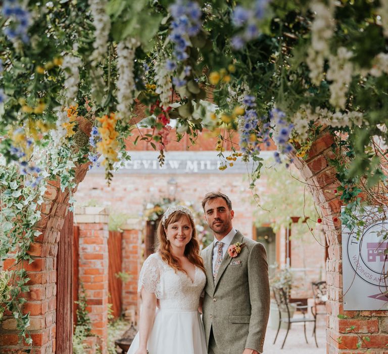 Groom in tweed suit with matching floral tie and pocket sqaure standing with the bride in a lace wedding dress with a white flower headband