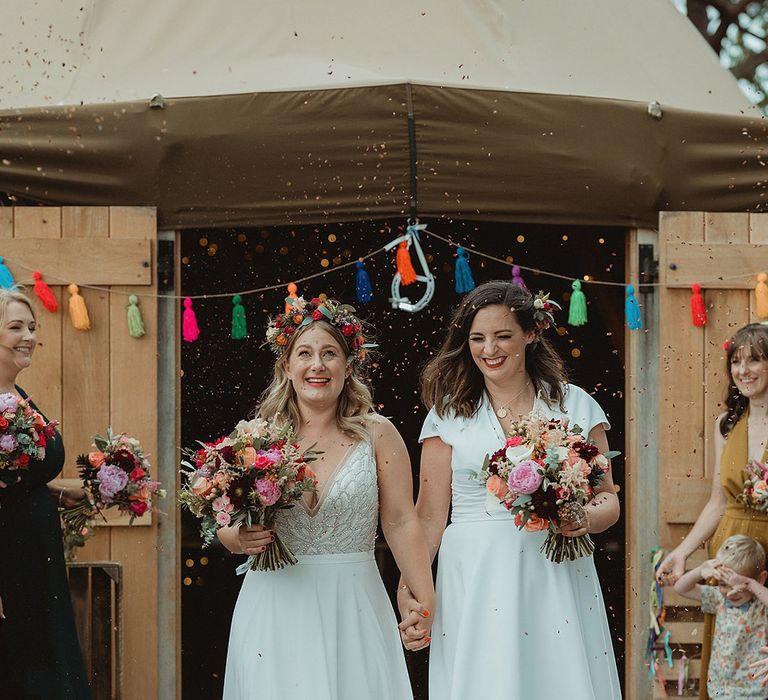 Bride exit their ceremony holding hands to a confetti exit wearing flowers in their hair and holding a colourful bouquet 