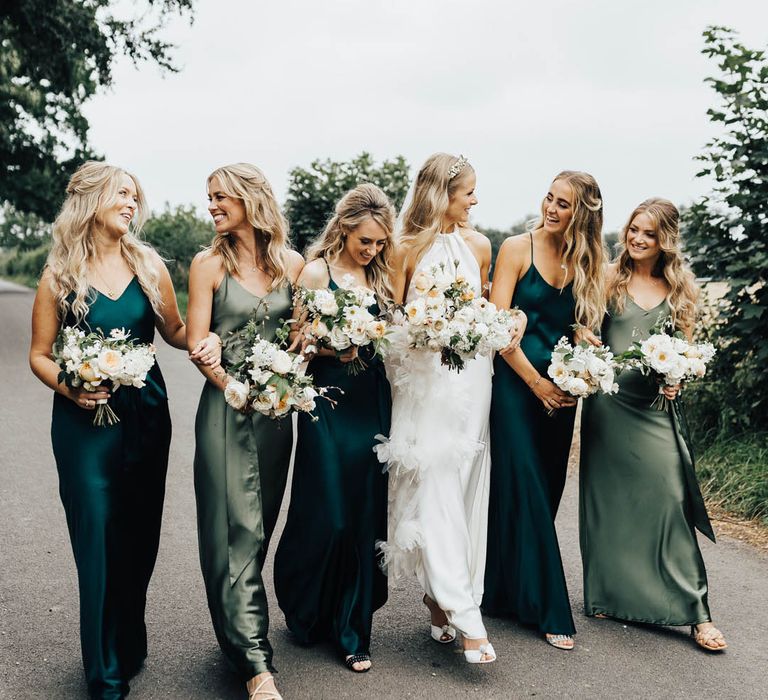 The bride in a ruffle feather wedding dress walks with the bridesmaids in satin dresses in mismatched shades of green