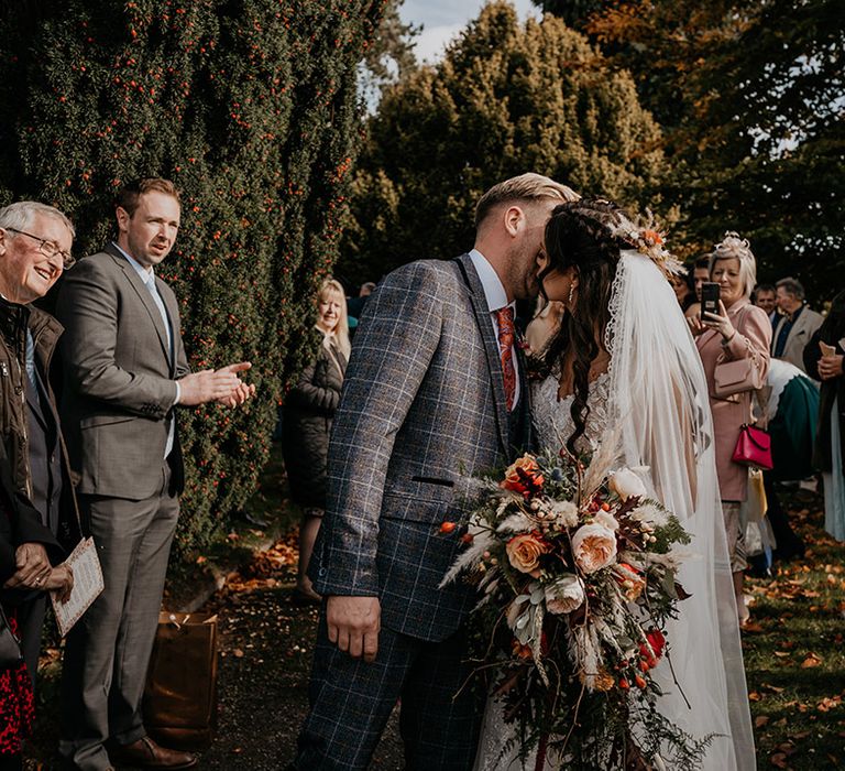 Groom in a grey checkered suit kisses the bride in a lace wedding dress holding a beautiful cascade wedding bouquet with dried grass as the wedding guests applaud