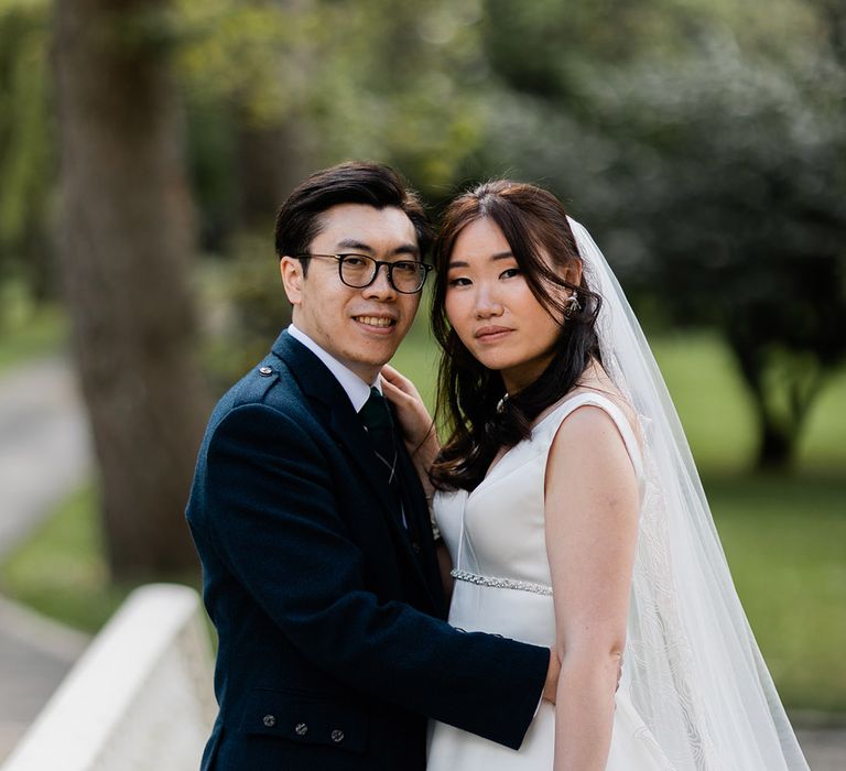 Bride wears her dark hair in loose curls and cathedral veil whilst stood beside her groom in green tartan kilt