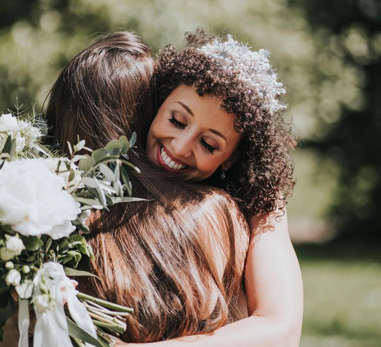 Bride wears embellished bridal crown in her naturally curly hair whilst hugging wedding guest 