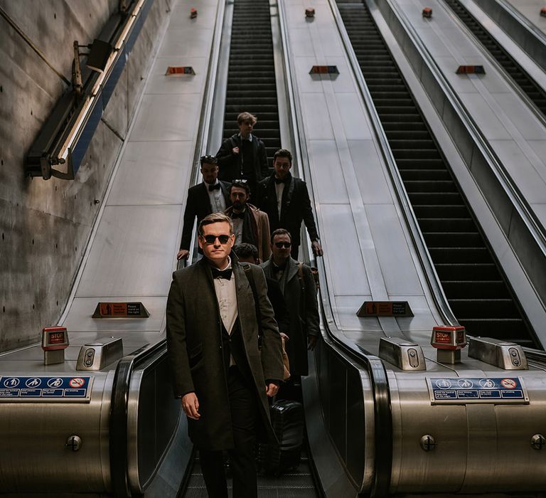 Grooms party in matching black tuxedos and sunglasses going down the escalator in the London Tube