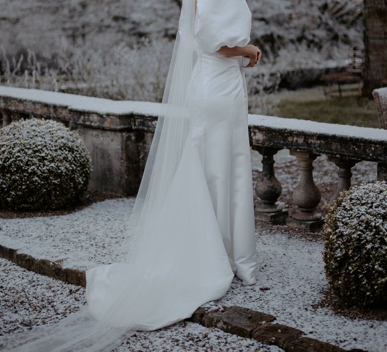 Bride in the grounds of snowy Eaves Hall Clitheroe wearing puff sleeve wedding dress with front slit and church