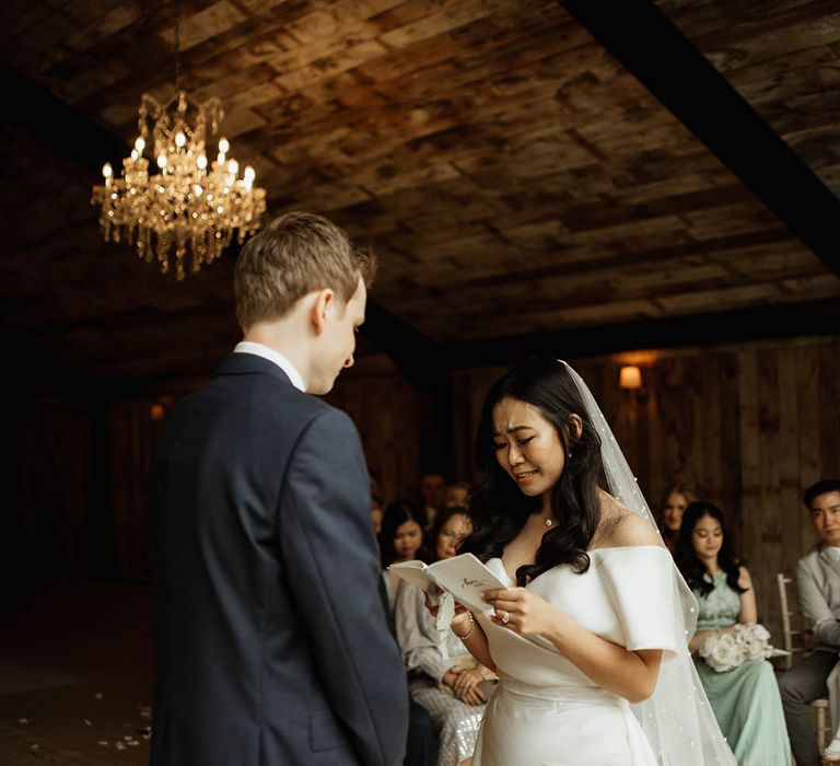 Bride gets emotional as she reads out her wedding vows in an off the shoulder wedding dress and pearl veil facing the groom in a navy suit for wedding at Hidden River Barns