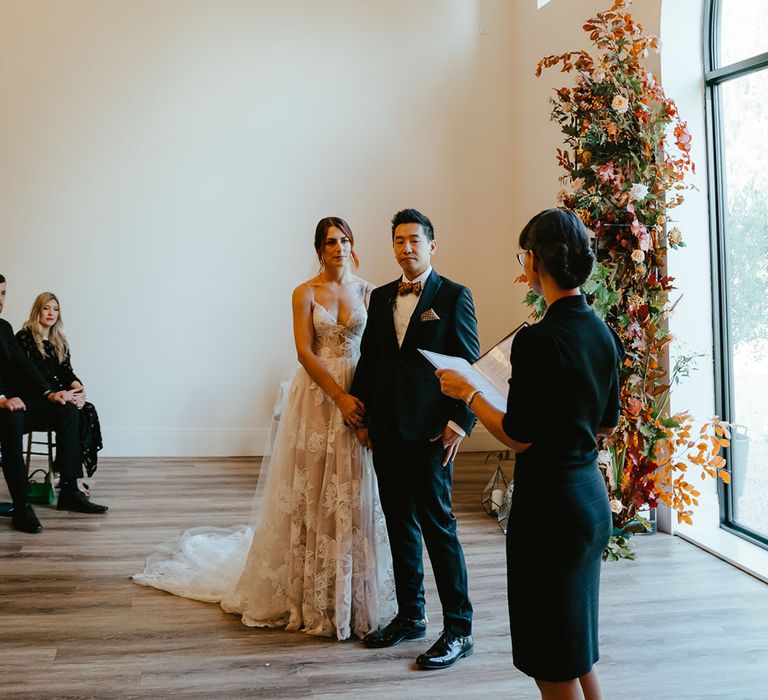 Bride & groom stand to the front of room beside burnt orange column flowers during wedding ceremony 