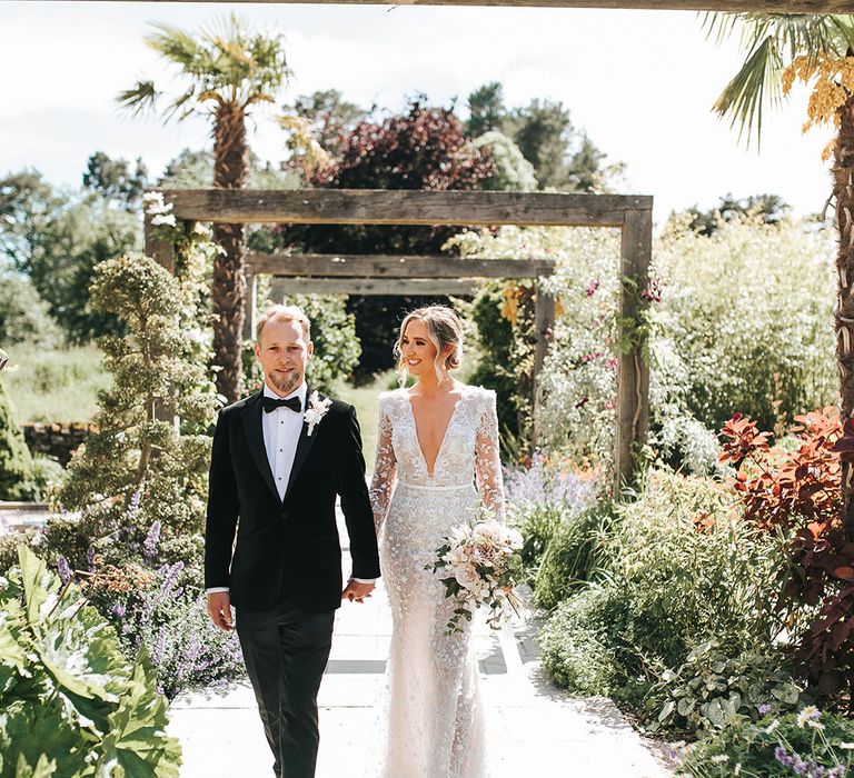 Groom in black tuxedo with white flower buttonhole walking with the bride in a pretty Wona wedding dress in the gardens at Le Petit Chateau in Northumberland