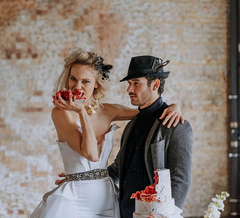 Bride eating red velvet wedding cake with her hands wearing romantic organza and tulle a-line wedding day bridal ballgown with a flowing skirt, sculpted bandeau top and patterned black belt and groom in dark grey jacket, black ribbed turtle neck, black trousers and a black fedora hat 