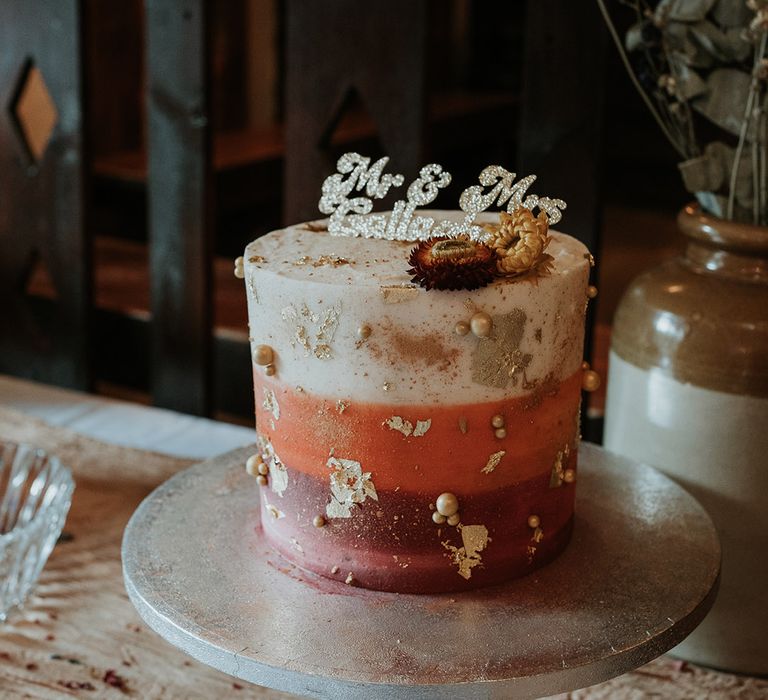 Single tier buttercream wedding cake in white, orange and pink on stone cake stand with personalised glittery wedding cake topper