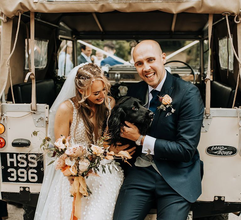 The groom sits in a navy suit and pink rose buttonhole with the pet dog and the bride in a Made With Love wedding dress after their confetti exit