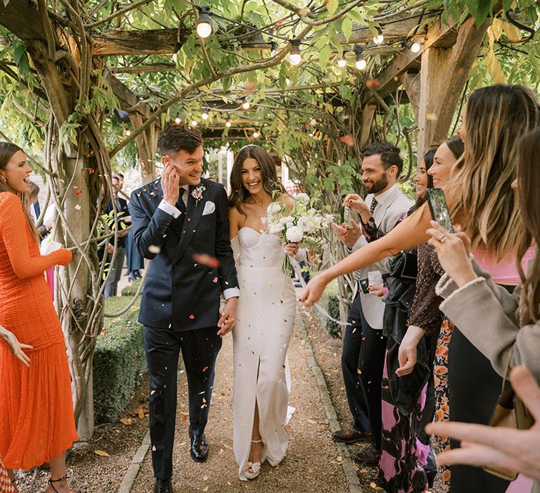 Groom in a dark suit walking with the bride in a fitted wedding dress with split leg to their confetti exit 