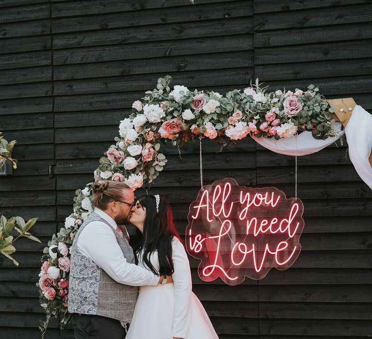 Neon pink sign reading 'All You Need Is Love' on a hexagonal wooden frame decorated with flowers with the bride and groom sharing a kiss next to it