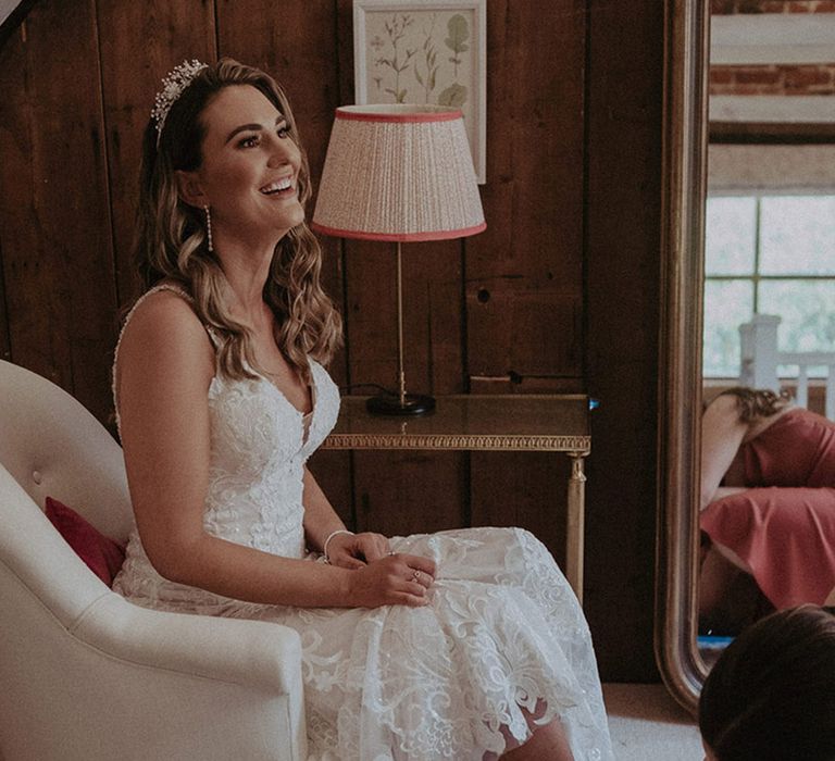 The bride sits in her fitted lace wedding dress with a tiara on as her bridesmaid helps her put on her pointed toe wedding shoes with a low heel