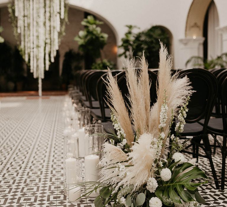 Tall white pillar candles in hurricane vases line the aisles with white dahlias and bell flower wedding flower and pampas grass arrangement 