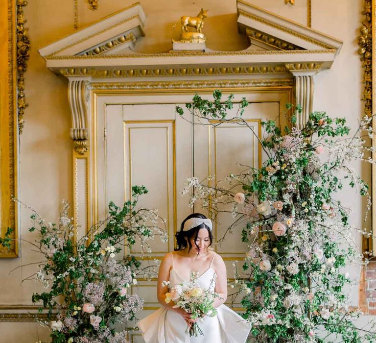 Bride in sleeveless satin wedding dress with large accent bow on the back standing in St Giles House with large rose, baby's-breath, foliage and dried flower floral columns holding classic spring bouquet 