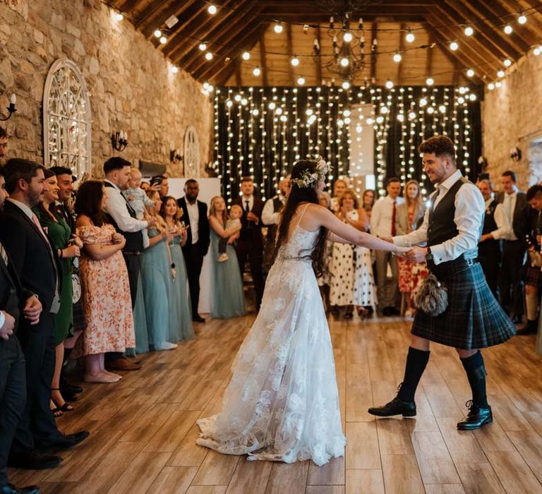 Bride in 3d applique floral sheer overlay wedding dress with white garden rose flower crown and cathedral-length veil dancing with groom in dark grey waistcoat with tartan tie and white garden rose boutonniere at The Barn at Harburn with suspended fairy lights and festoon lighting 