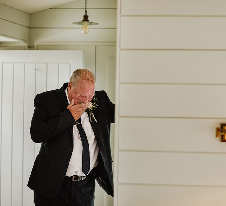 Father of the bride in black tie with gypsophila buttonhole cries as he sees the bride in her wedding dress for the first time 