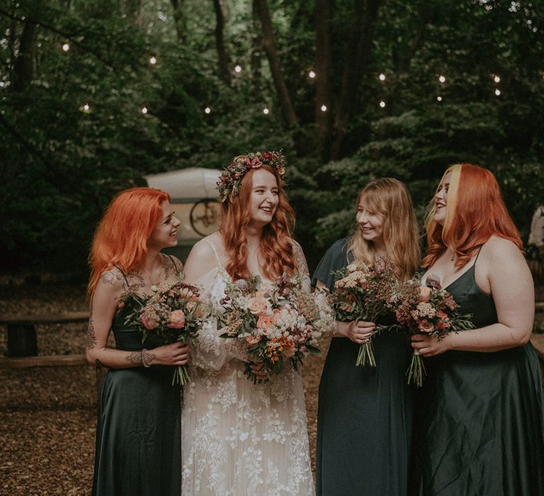 Bride in off the shoulder boho lace wedding dress and pink flower crown with the bridesmaids in green satin bridesmaid dresses 