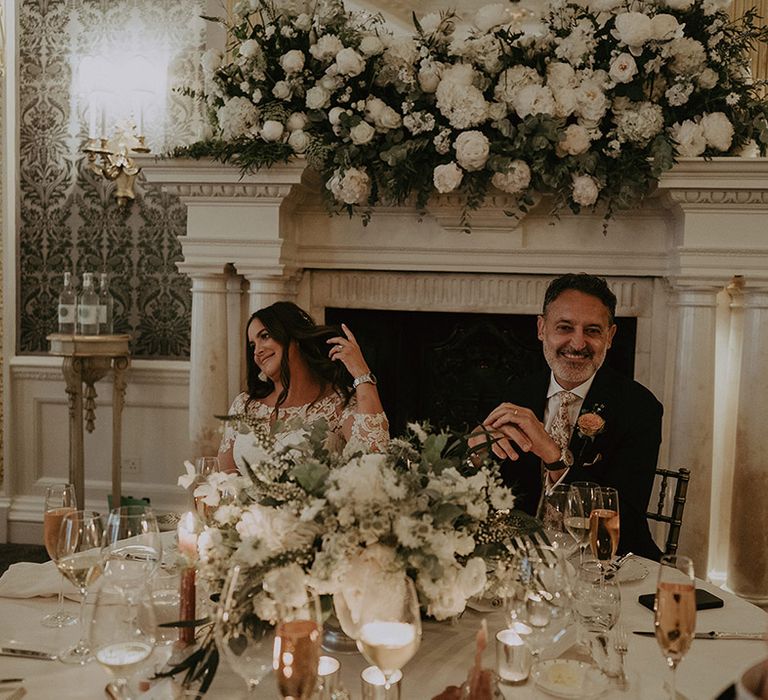 The bride and groom sit at their round table with white tablecloth with pink and white floral arrangements 