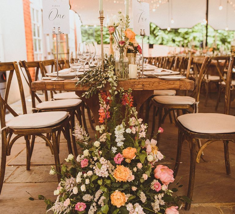 Long banquet tables decorate dight orange, pink, and white wedding floral arrangements with table number signs and pastel taper candles 