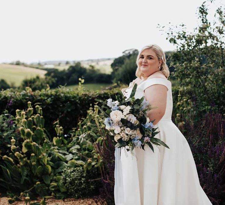 Bride standing on the grounds of Brickhouse Vineyard wedding venue wearing satin off the shoulder wedding dress holding white and blue eucalyptus, foliage, garden rose, gardenia, light blue delphinium and dried flower bridal bouquet tied with dusky blue ribbon 