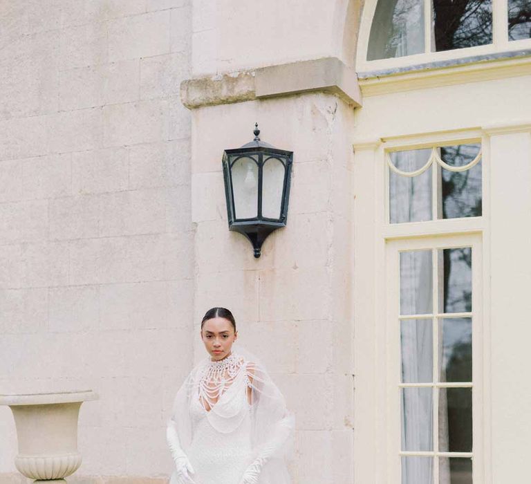 Bride wearing low v-neck slip bridal dress with dramatic pearl necklace and accessories, detachable tulle sleeves and tulle puddle train and white satin wedding gloves at Settrington Orangery