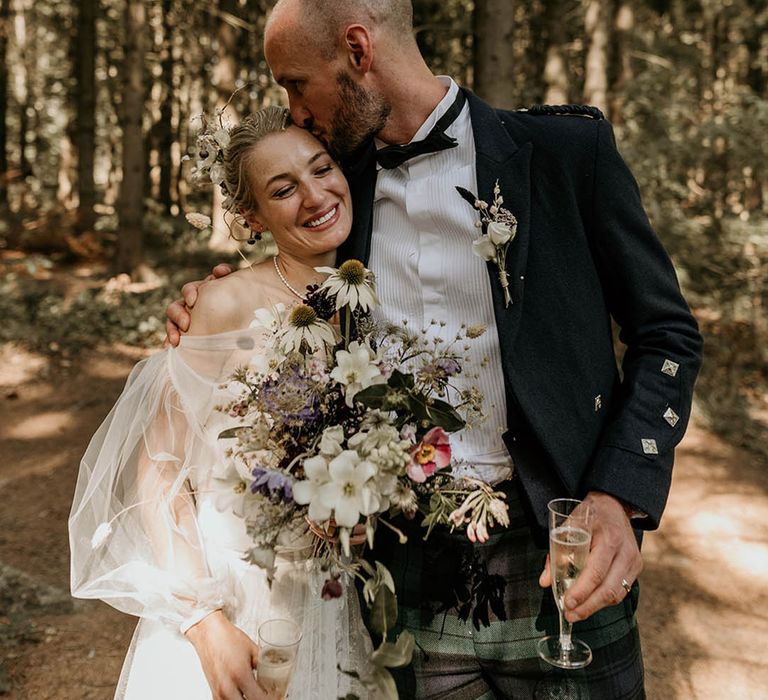 Groom in bespoke checkered trousers and pleated shirt kissing bride on the forehead holding a wildflower wedding bouquet 