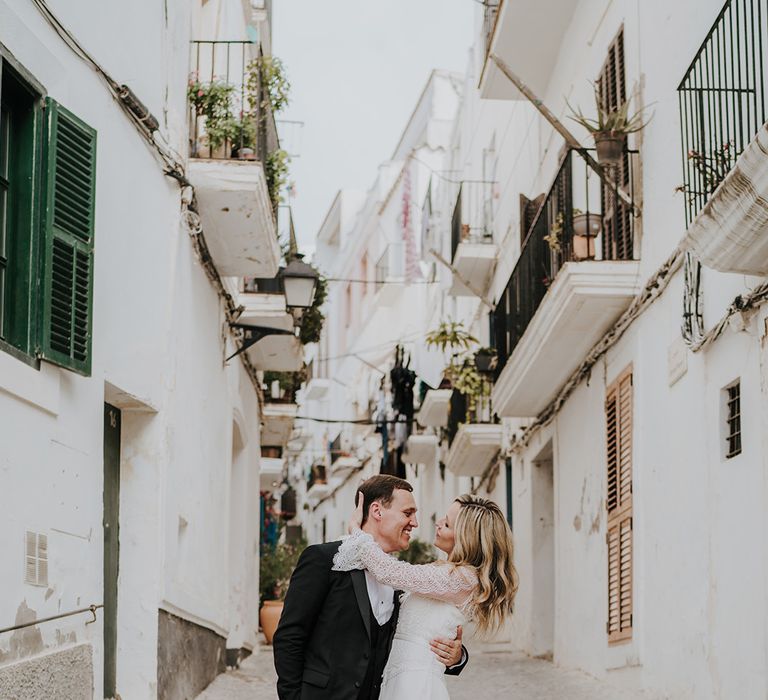 bride in a boho lace wedding dress with long sleeves cupping her grooms head in the side streets of ibiza with white buildings balconies and shutters