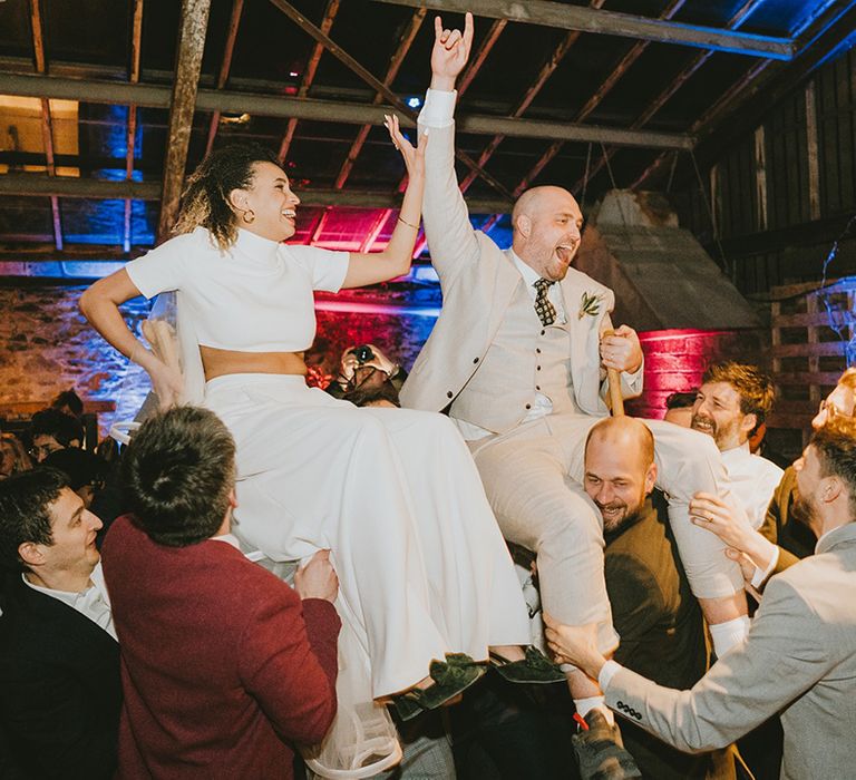 The bride in a short sleeve cropped top and trousers by Jesus Peiro being lifted by the wedding guests next to the groom in a cream suit