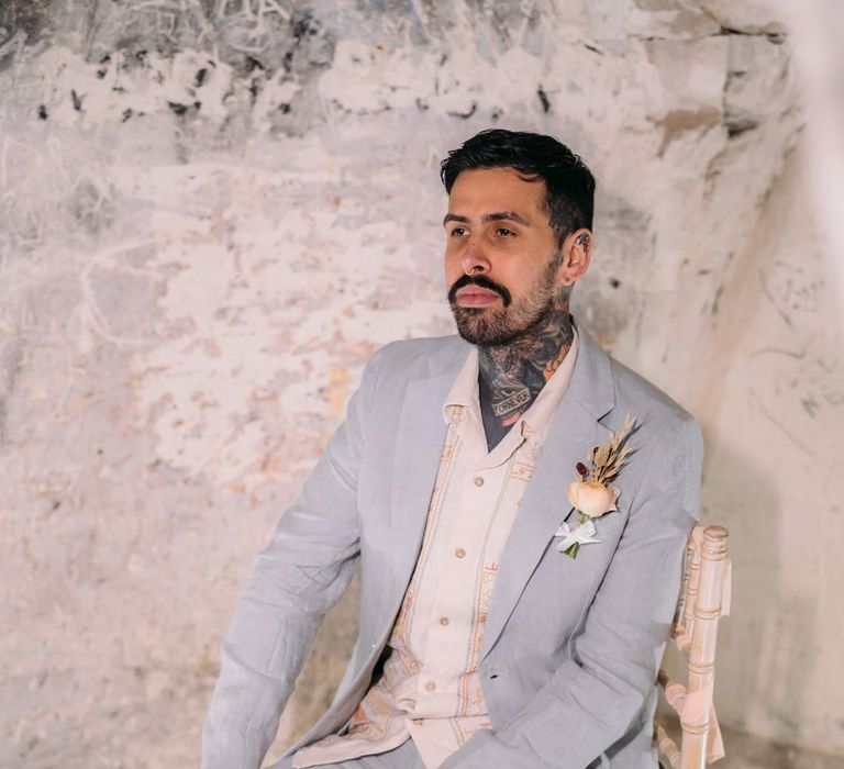 Groom in baby blue linen suit, personalised embroidered linen shirt and garden rose and pampas grass boutonniere sitting on chair in Margate Caves