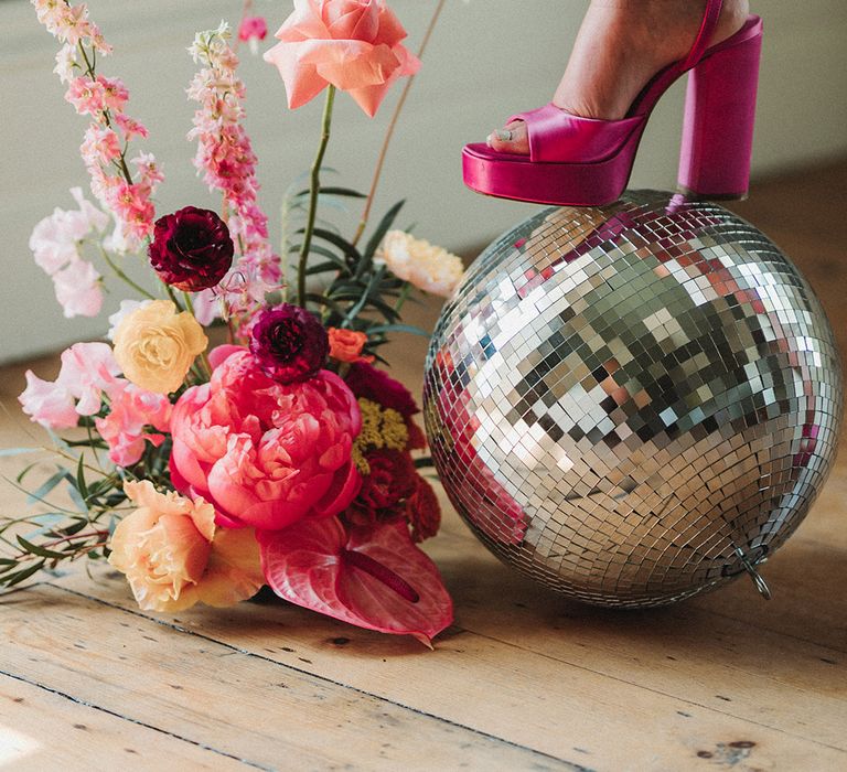 Bride in bold statement hot pink platform shoes resting on disco ball with pink wedding flower arrangements 